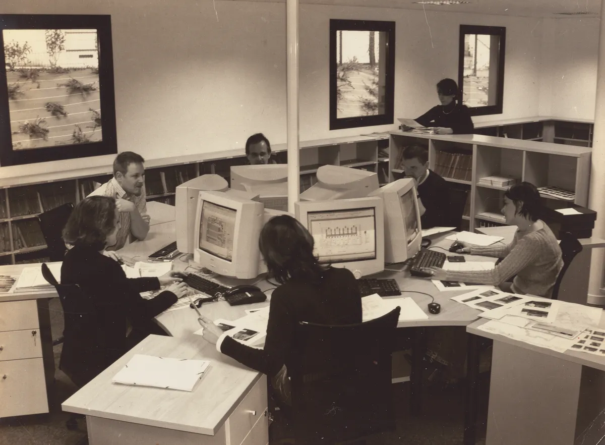 Photographie sépia du bureau d’études de SIB : des collaborateurs travaillent sur des écrans cathodiques, entourés de plans et de planches de photographies de relevés.